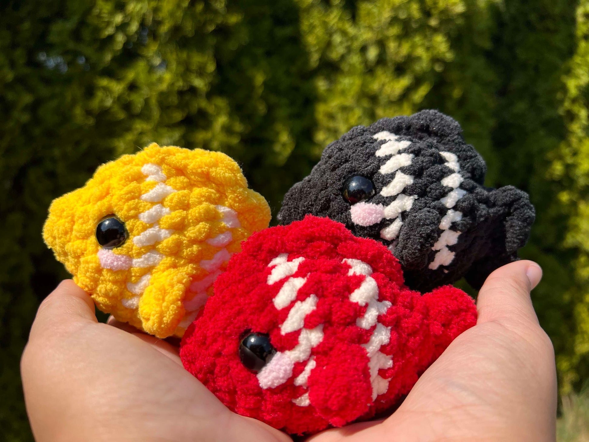 Group of 3 crochet plushie fishes being held with trees in the background.